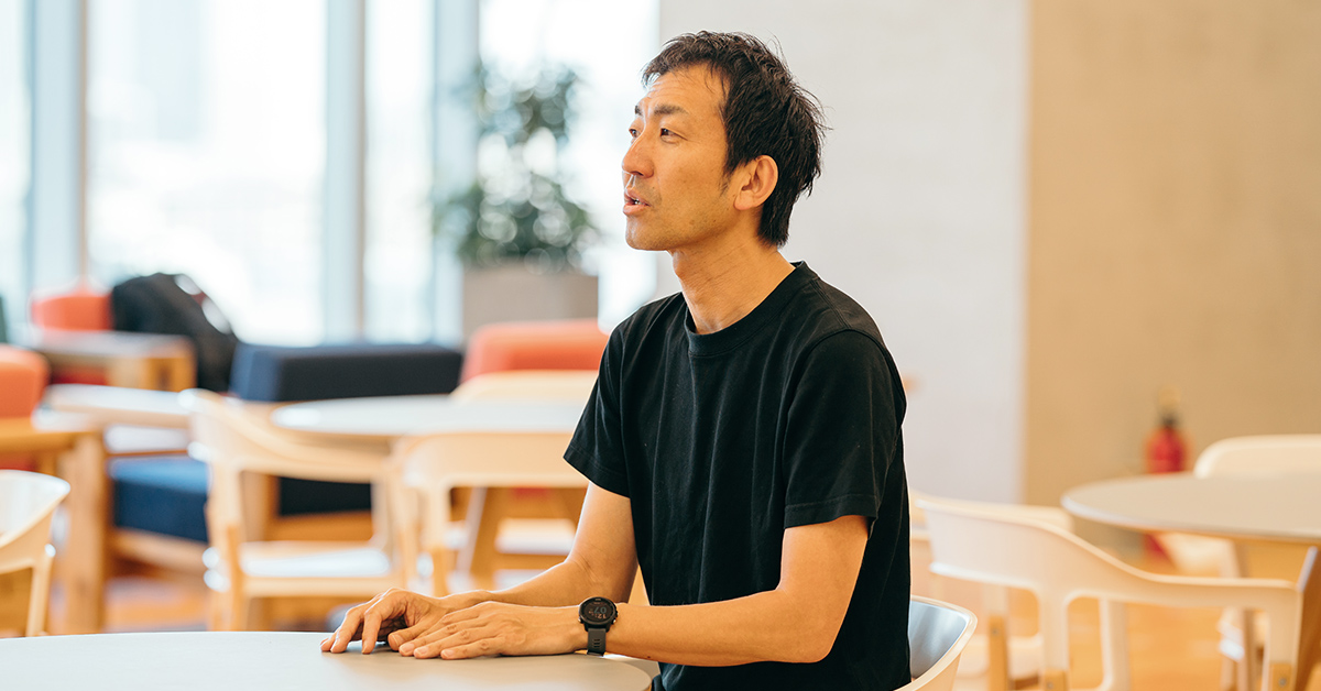 カフェのような場所で、黒いシャツの男性が座って話しています。