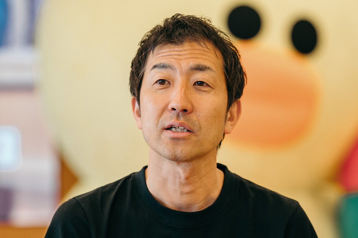 黒いシャツの男性が、大きなキャラクターの前で話しています。