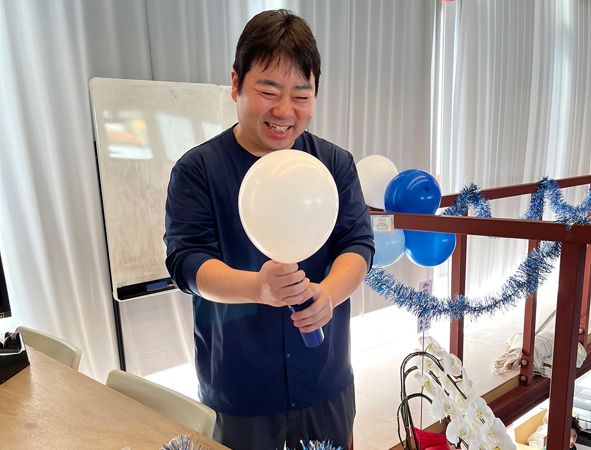 田宮が白い風船を持って微笑んでいます。背景には青と白の風船や飾り付けがあり、楽しげな雰囲気です。