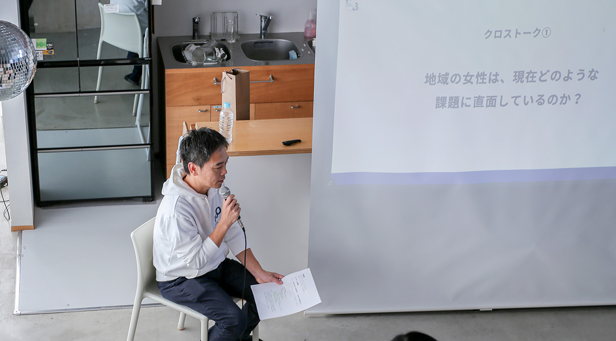 椅子に座った和田さんがマイクを持ち、プレゼンテーションを行っています。スクリーンには「クロストーク① 地域の女性は、現在どのような課題に直面しているのか？」と表示されています。