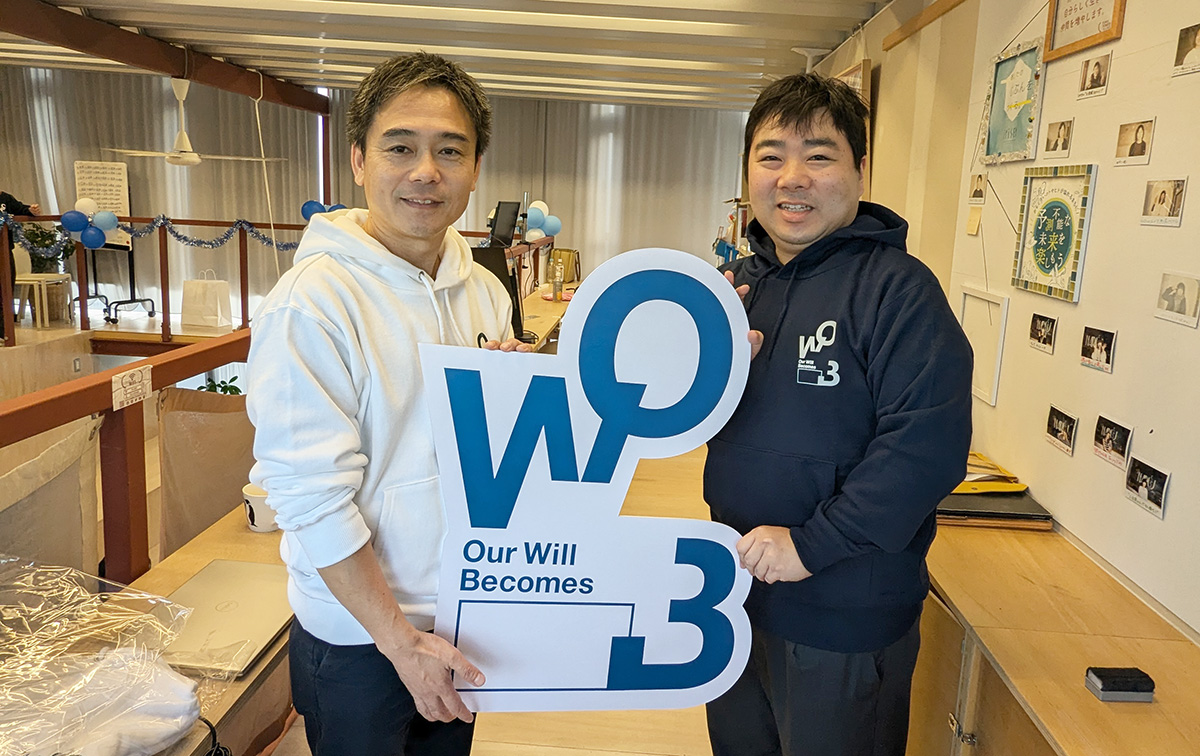 和田さんと田宮が「W03 Our Will Becomes」という大きなサインを持ち、微笑んでカメラを見ています。背景には飾り付けがされています。
