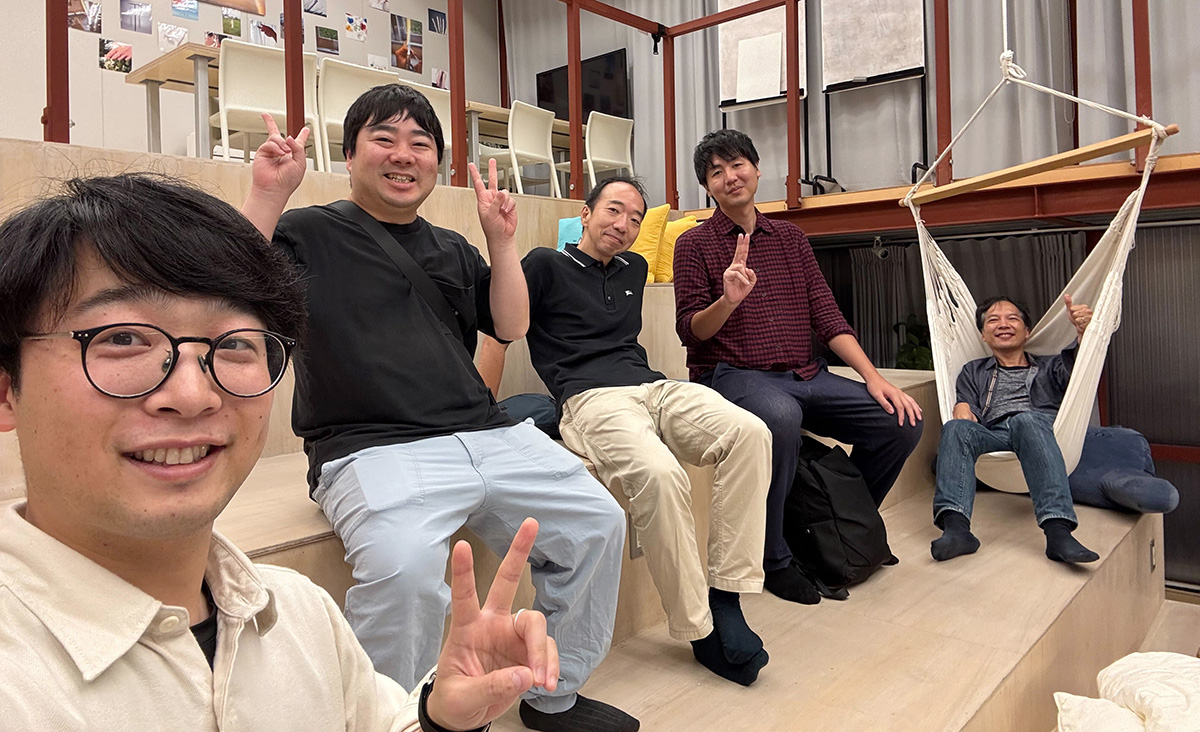 LINEヤフーの5人の男性社員が階段状の座席に座り、ピースサインをして笑顔でカメラを見ています。右端の男性はハンモックに座っています。楽しげな雰囲気です。