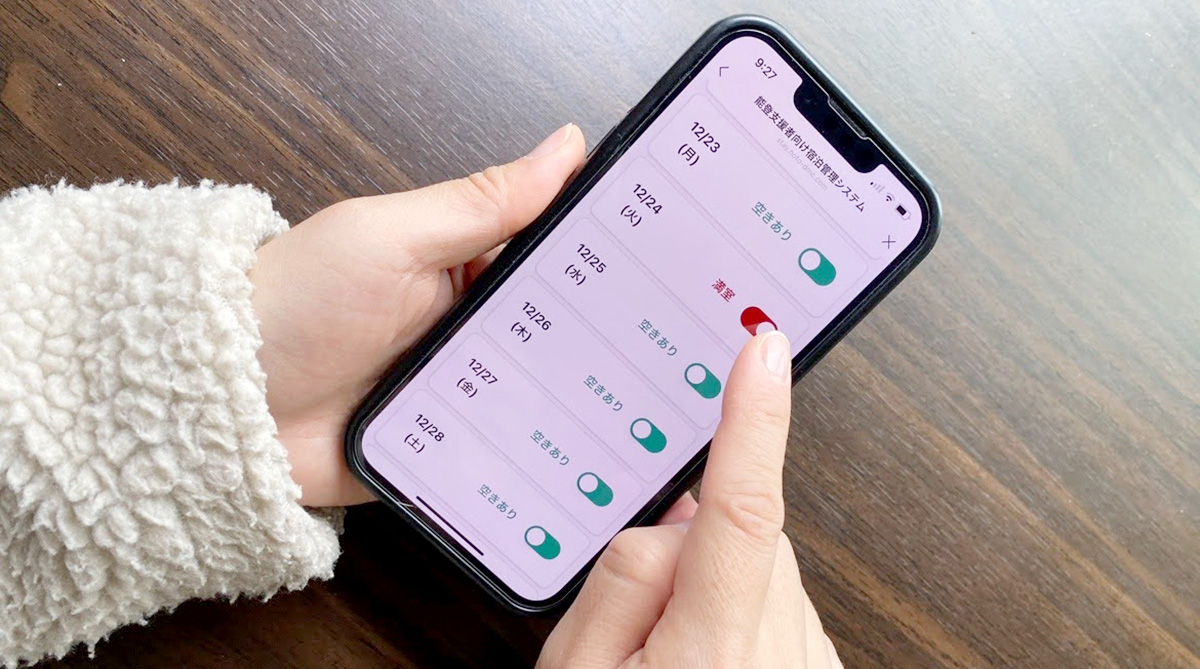 スマートフォンの画面に、日付ごとの空室状況が表示されています。指が「満室」のスイッチを確認している様子です。