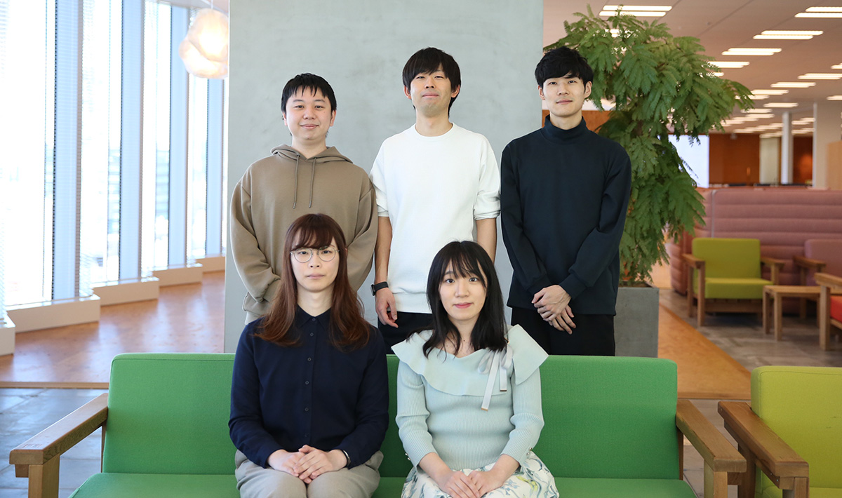 5人の人物がオフィスのソファに座り、カメラに向かって微笑んでいる集合写真です。