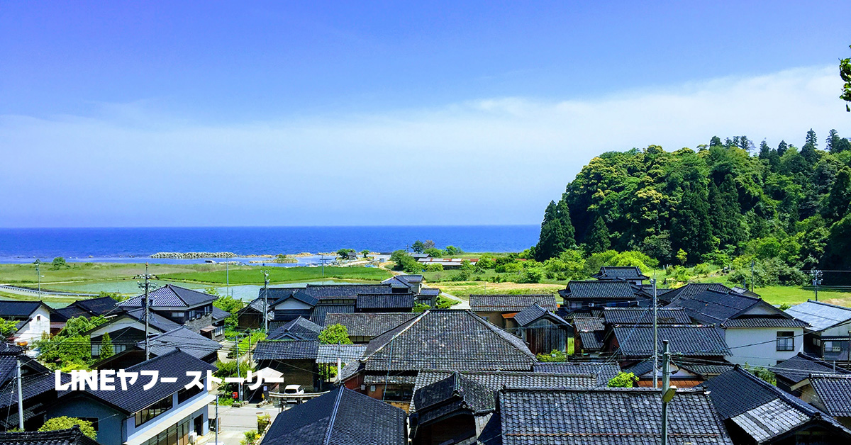 能登の風景で、黒い瓦屋根の家々と青い海が広がっています。緑豊かな山々が背景にあり、「LINEヤフー ストーリー」と書かれています。