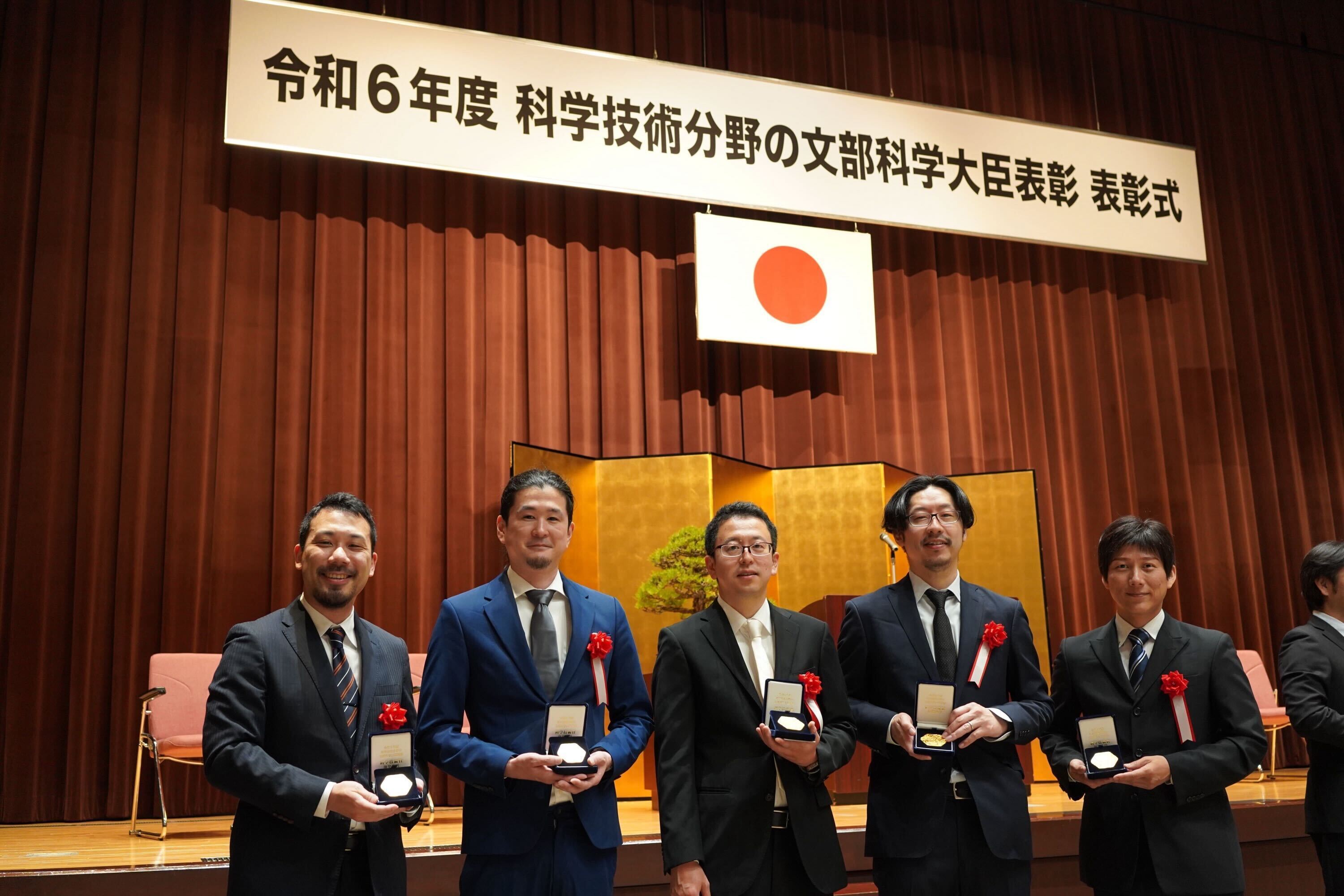 表彰式で5人の男性がメダルを持ち並んでいる写真。