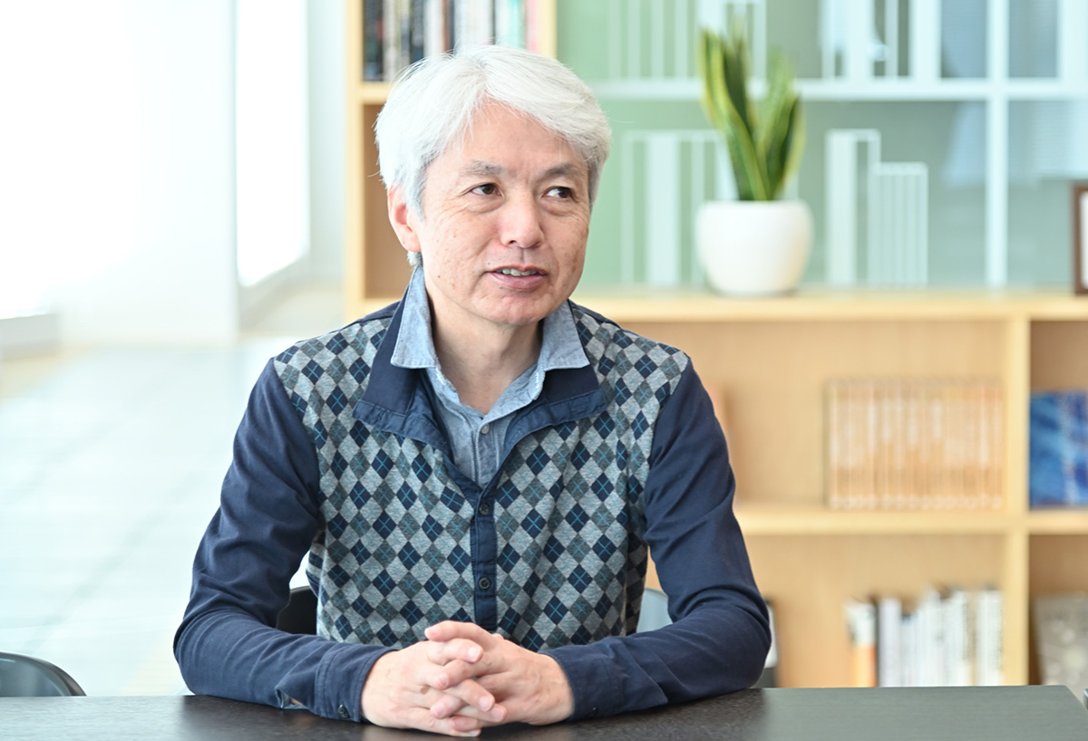 岩崎がオフィスのテーブルに座り、話をしている様子です。背景には本棚と観葉植物があります。