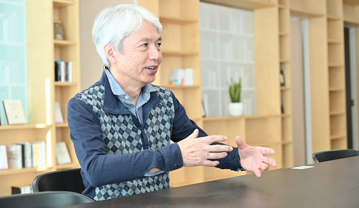岩崎がオフィスのテーブルで話をしている様子です。背景には本棚と観葉植物が見えます。