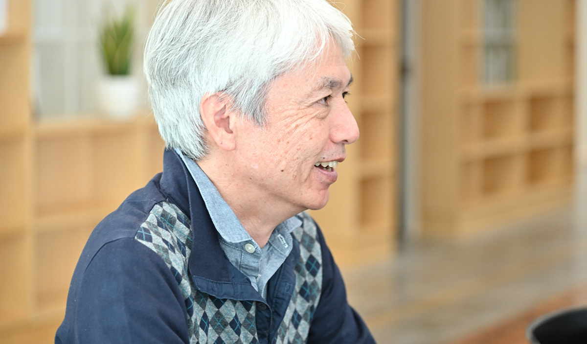 岩崎がオフィスのテーブルで話をしている様子です。背景には本棚と観葉植物が見えます。