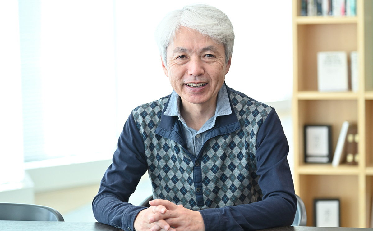 岩崎がオフィスのテーブルに座り、笑顔でカメラを見ています。背景には本棚があり、明るい雰囲気です。