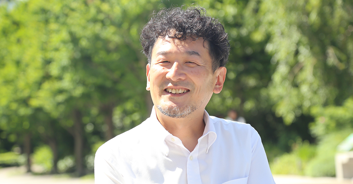 A man in a white shirt smiles against a lush, green background.