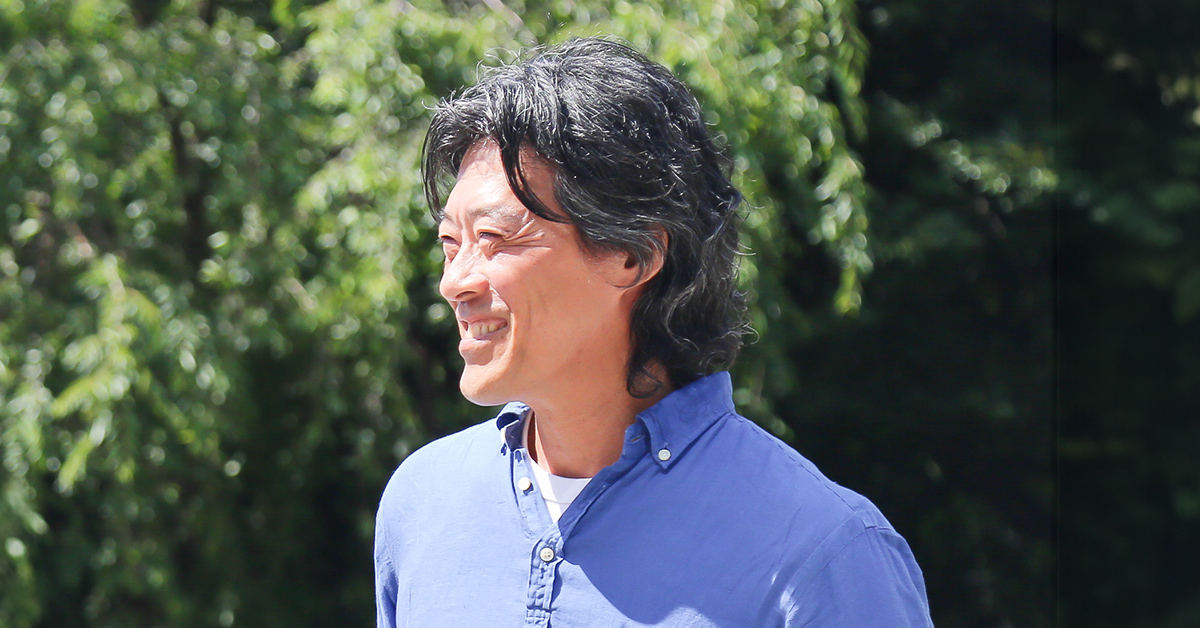 A man in a blue shirt smiles, appearing relaxed in a natural setting.