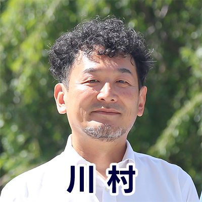 男性が屋外でカメラに向かって微笑んでいる。背景には緑が広がり、名前が「川村」と表示されている。