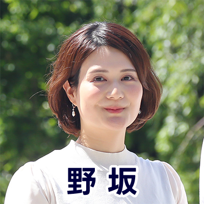 女性が屋外でカメラに向かって微笑んでいる。背景には緑が広がり、名前が「野坂」と表示されている。