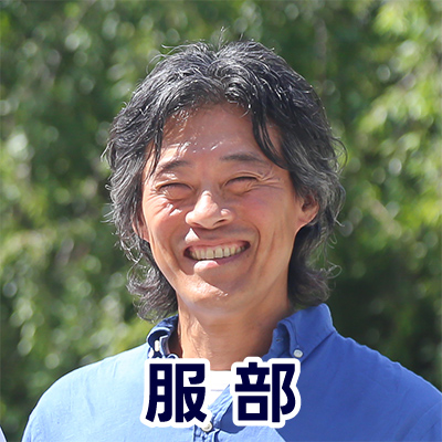 男性が屋外でカメラに向かって笑顔を見せている。背景には緑が広がり、名前が「服部」と表示されている。