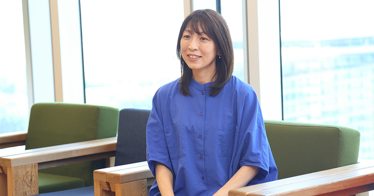 女性が窓際の椅子に座っている。青い服を着ており、リラックスした様子で微笑んでいる。