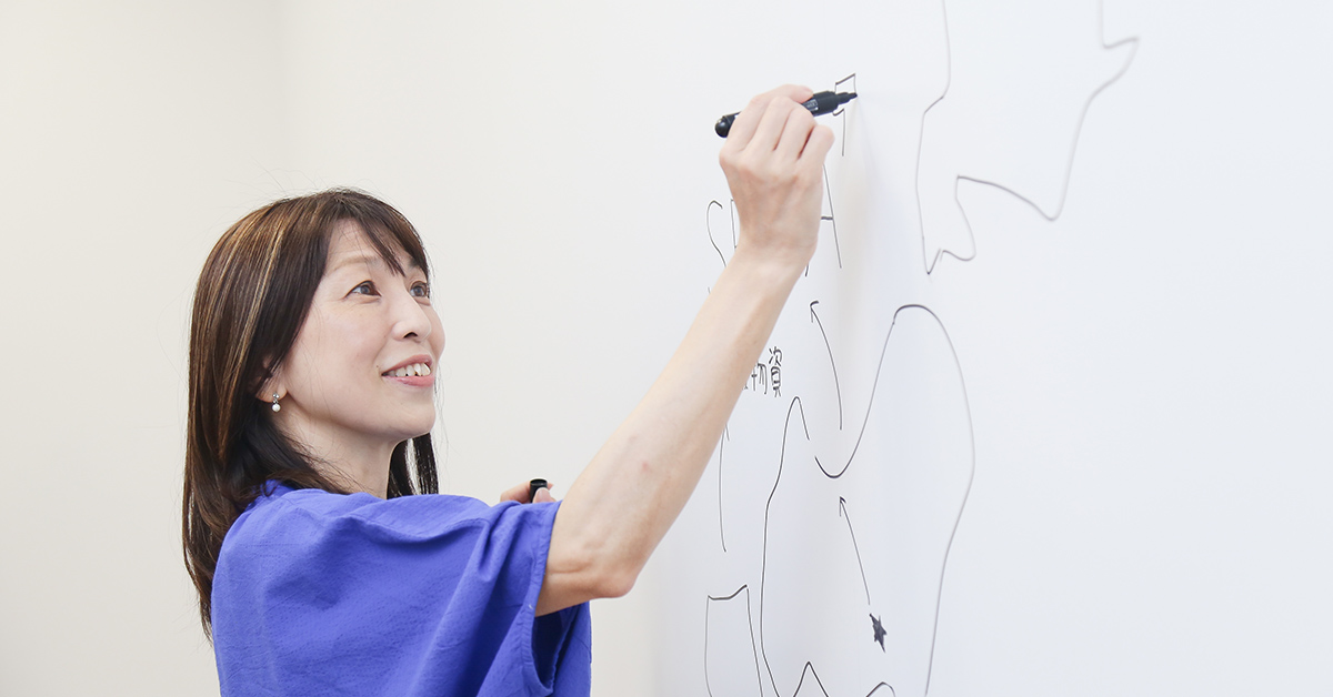 女性がホワイトボードに図を描いている。青い服を着ており、集中した表情をしている。
