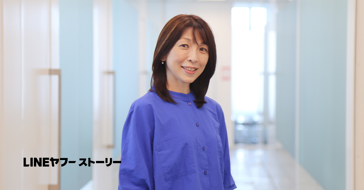 女性が廊下に立ち、微笑んでいる。青い服を着ており、背景には明るい光が差し込んでいる。