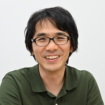 メガネをかけた男性が、緑色のシャツを着て笑顔でカメラを見ています。背景はシンプルな白い壁です。