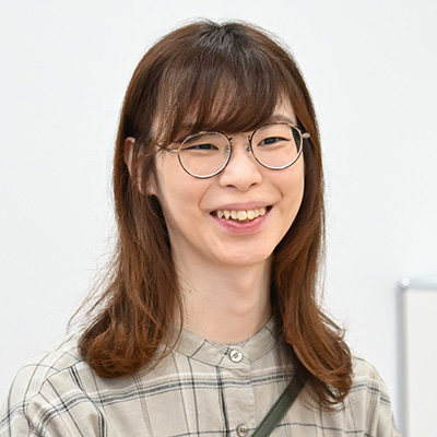 メガネをかけた女性が、チェック柄のシャツを着て微笑んでいます。背景は白い壁でシンプルです。