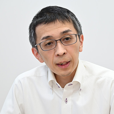 メガネをかけた男性が白いシャツを着て、話をしている様子です。背景は白い壁でシンプルです。