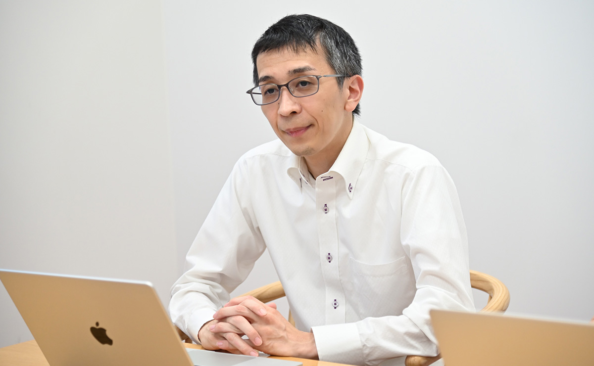 白いシャツを着た男性が、ノートパソコンの前で話をしています。背景はシンプルな室内です。