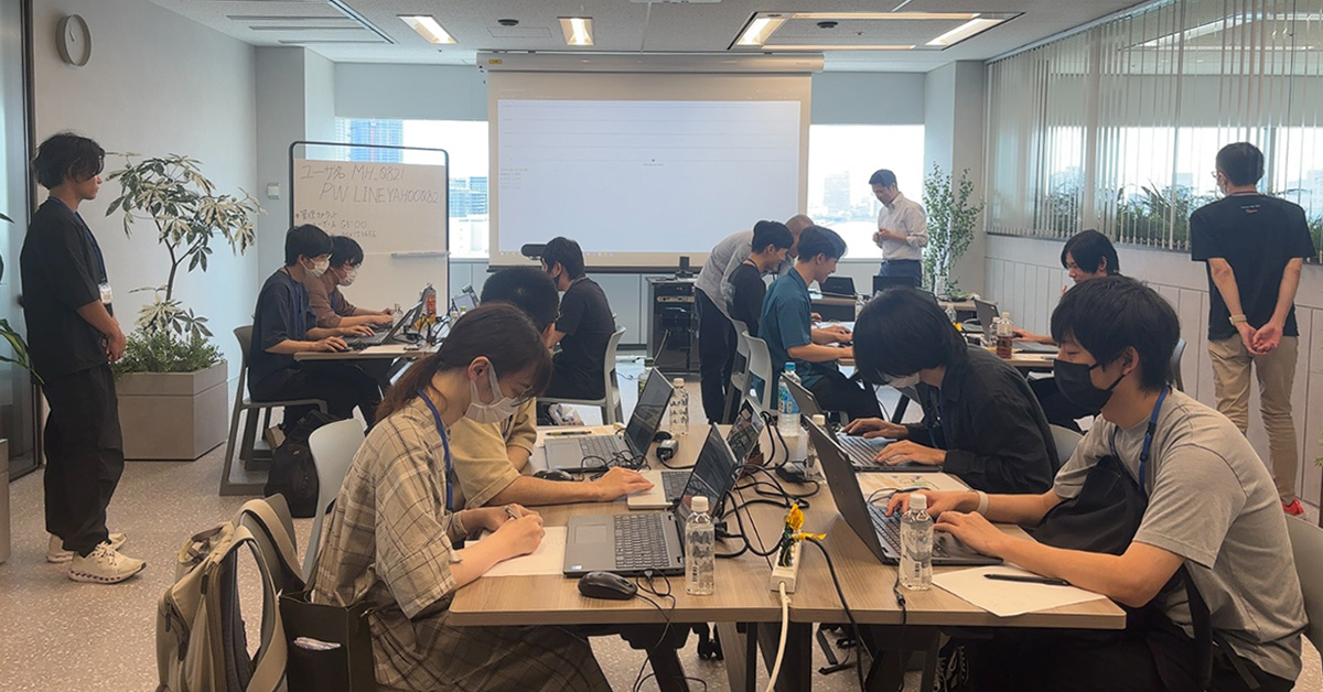 会議室で人々がノートパソコンを使って作業をしています。背景にはホワイトボードとプロジェクターがあります。