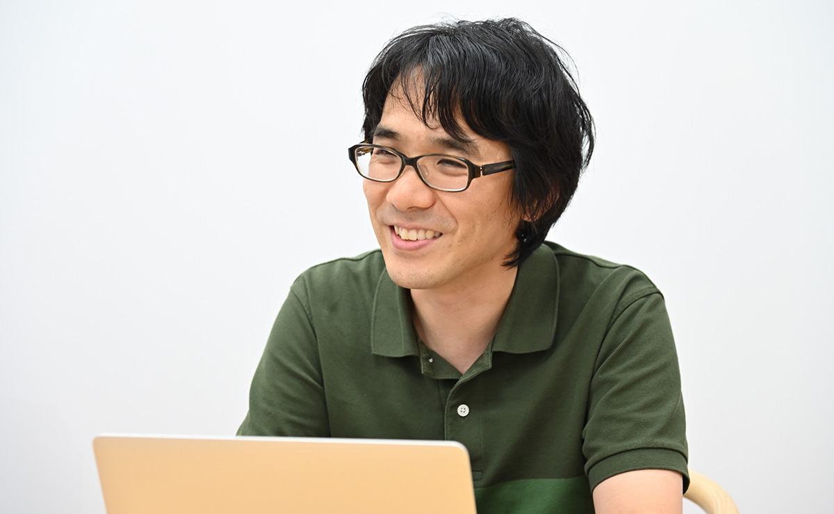 緑色のシャツを着た男性が、ノートパソコンの前で笑顔で話をしています。背景はシンプルな室内です。