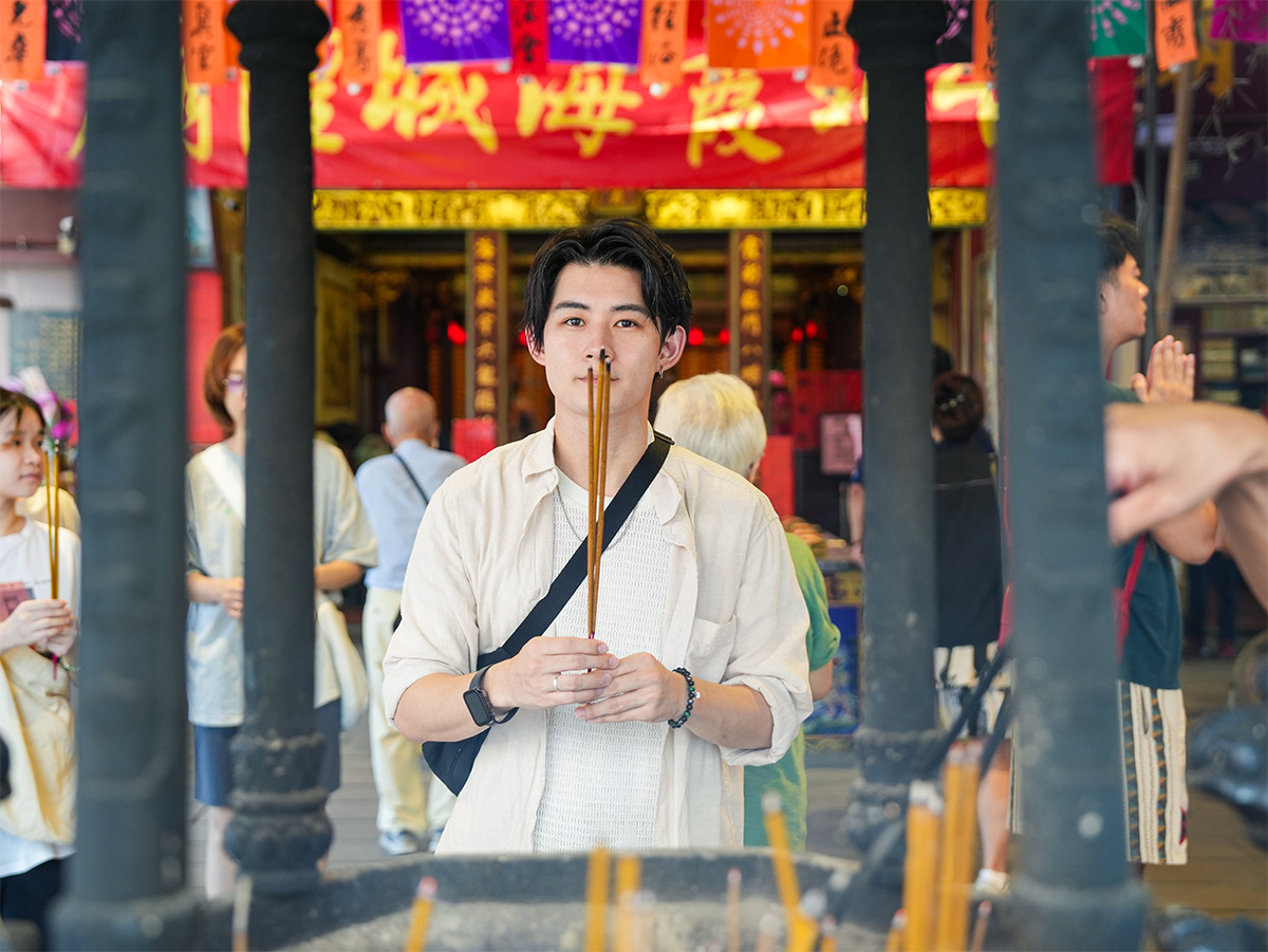 男性が寺院で線香を持っている写真です。背景には多くの参拝者が見えます。