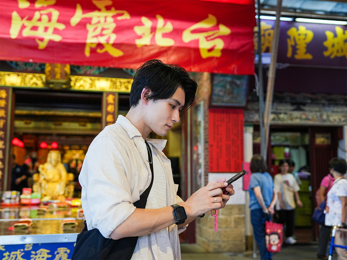 男性がスマートフォンを操作している写真です。背景には赤い看板と多くの人々が見える賑やかな場所です。