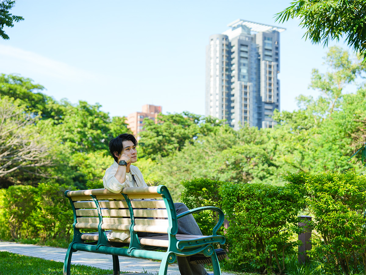 男性が公園のベンチに座り、遠くを見つめている写真です。背景には高層ビルが見えます。