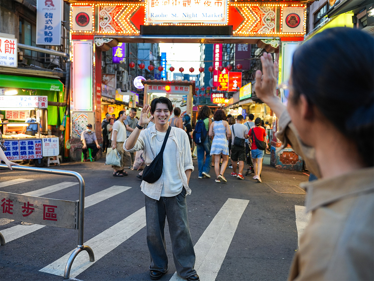 夜市の入り口で、一人の男性が手を振って挨拶しています。背景には多くの人々が歩いています。