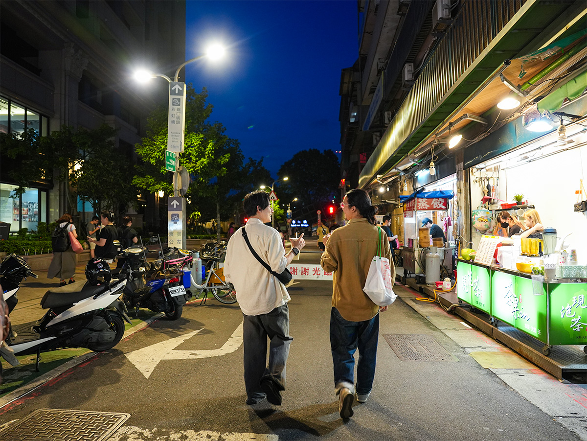 二人の男性が夜の通りを歩いています。屋台が並び、賑やかな雰囲気が漂っています。