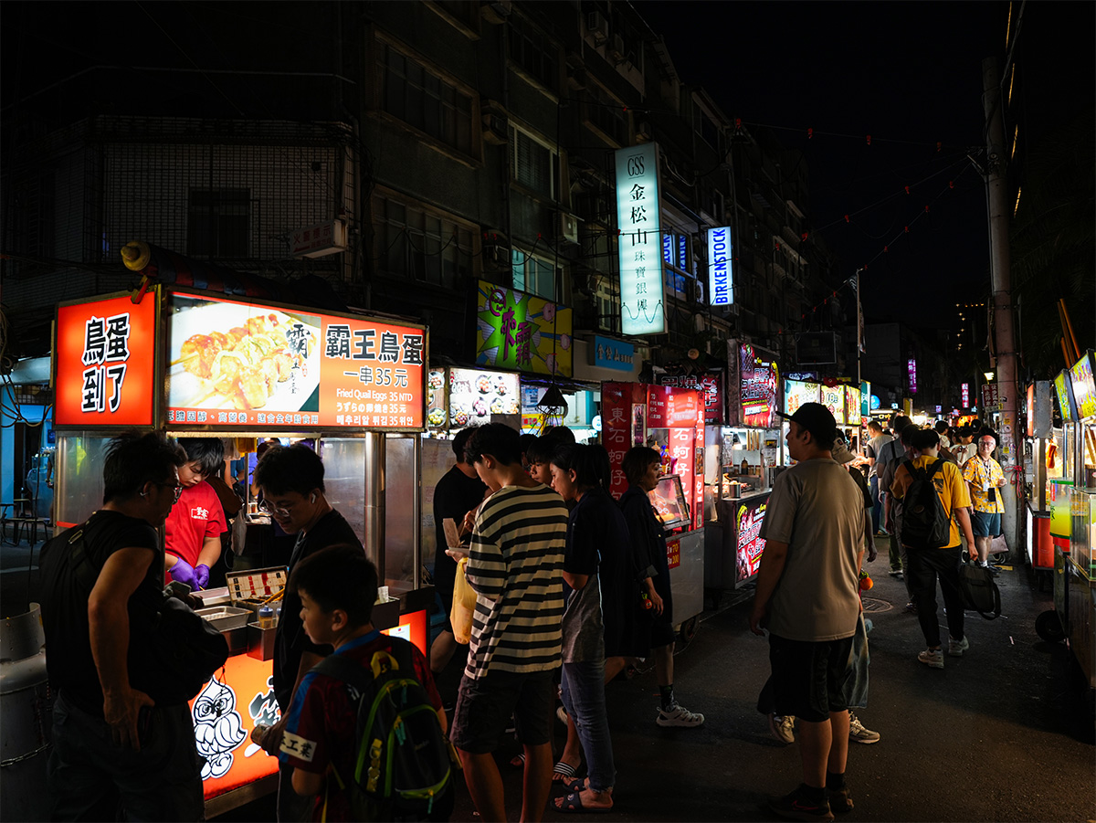 夜市の屋台が並び、多くの人々が食べ物を楽しんでいます。色鮮やかな看板が目立ちます。