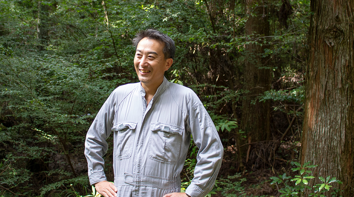 森の中で笑顔の男性が立っています。彼はグレーの作業服を着て、楽しそうにしています。
