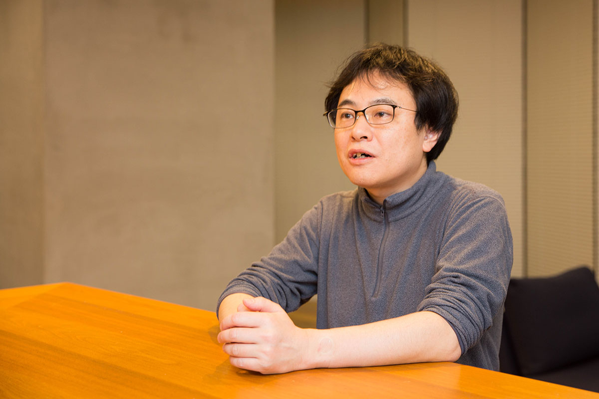 眼鏡をかけた男性が、机に手を置いて話している写真です。カジュアルな服装で、室内の落ち着いた雰囲気が感じられます。