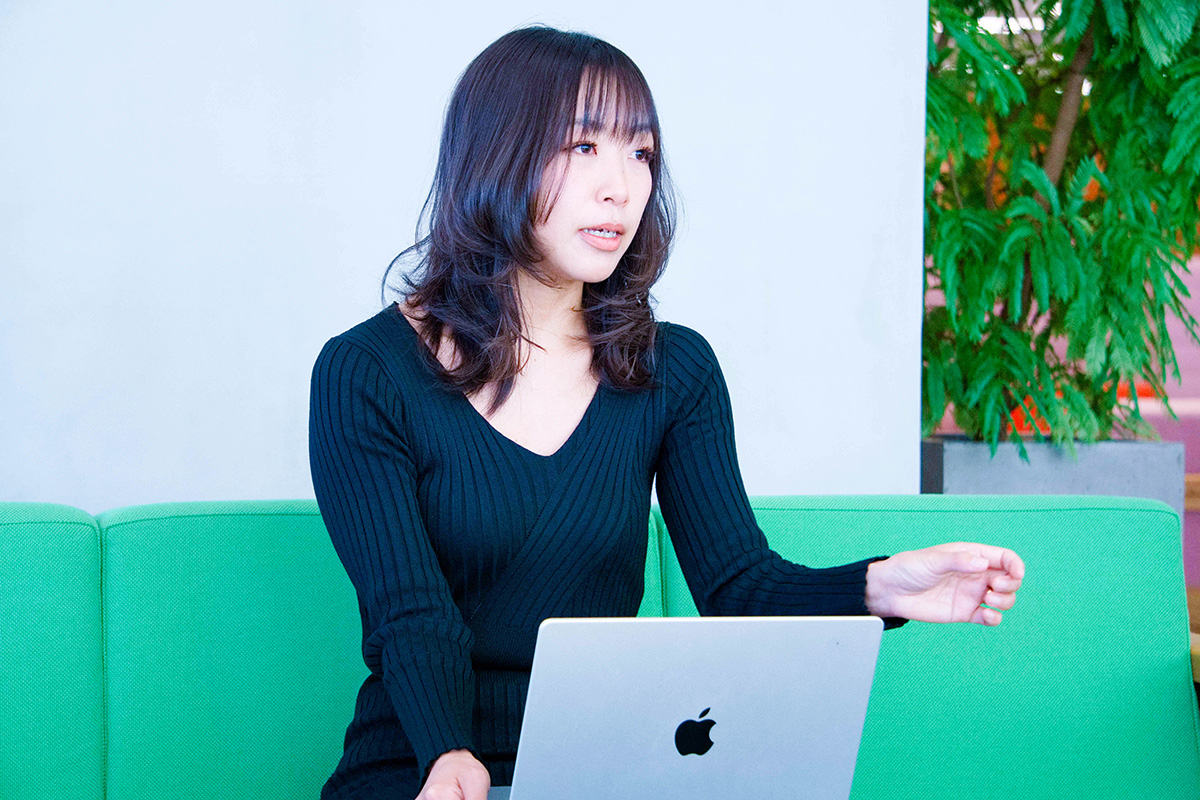 女性が緑のソファに座り、ノートパソコンを前にして話しています。背景には植物があり、リラックスした雰囲気です。