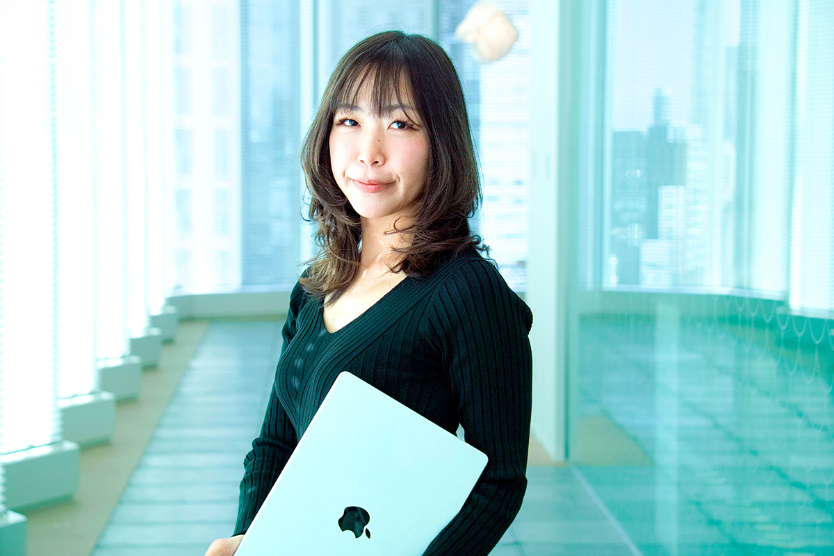 女性がノートパソコンを抱え、明るい廊下で微笑んでいます。背景には都会的な景色が広がっています。
