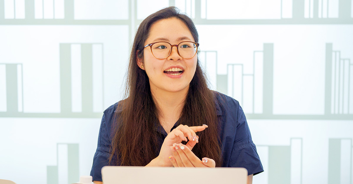 女性がノートパソコンの前で話をしている。背景には本棚のようなデザインがある。