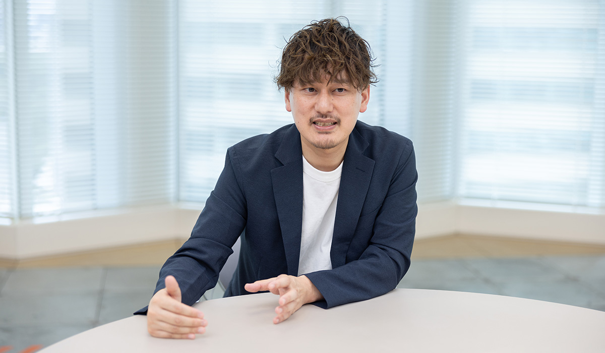 男性がテーブルに座り、手を動かしながら話している。背景には大きな窓が見える。