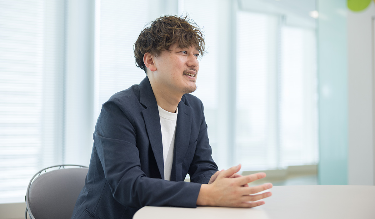 男性が横を向いて座り、手を組みながら話している。明るい室内でのシーン。