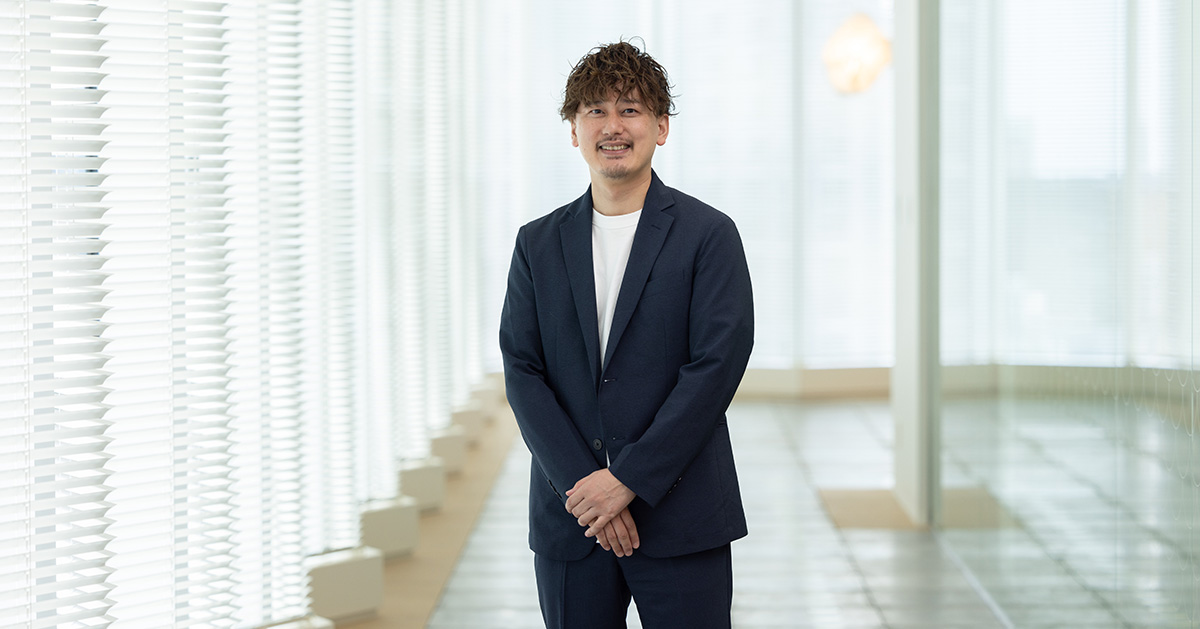 男性が廊下に立ち、手を前で組んでいる。明るい窓が背景に広がっている。
