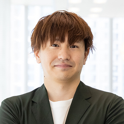 男性が白い背景の前で微笑んでいる写真です。彼はカジュアルな服装をしています。