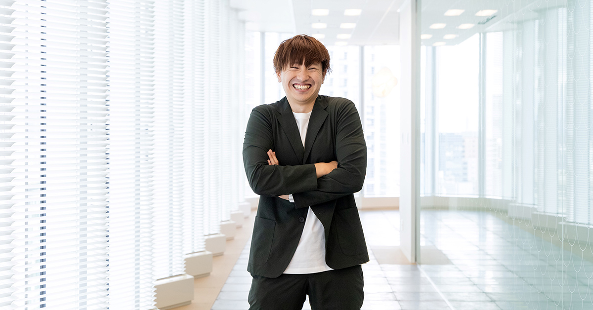 男性が明るい廊下で腕を組んで立っている写真です。彼はリラックスした様子で笑顔を見せています。