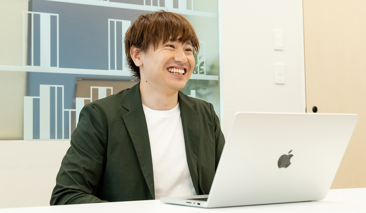 男性がノートパソコンの前で笑顔を見せている写真です。背景には幾何学的なデザインが見えます。