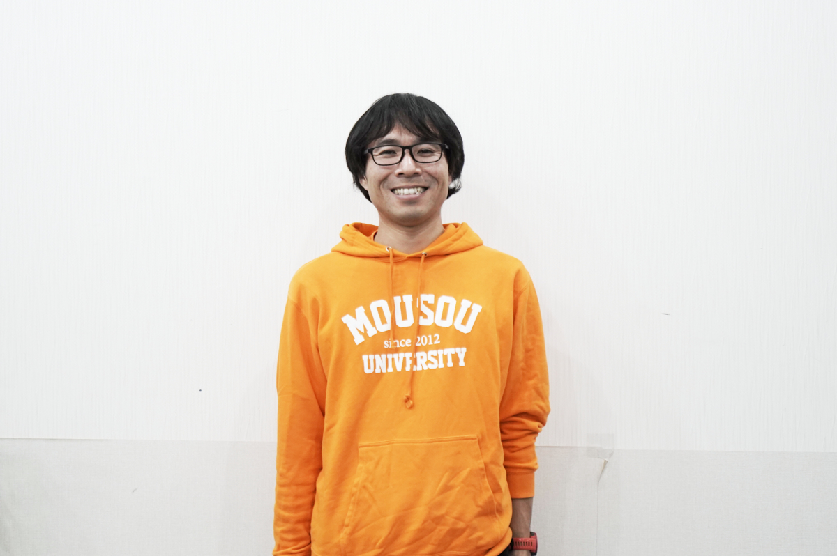 オレンジ色のパーカーを着た男性、地主さんが笑顔で立っています。パーカーには「MOUSOU UNIVERSITY」と書かれています。