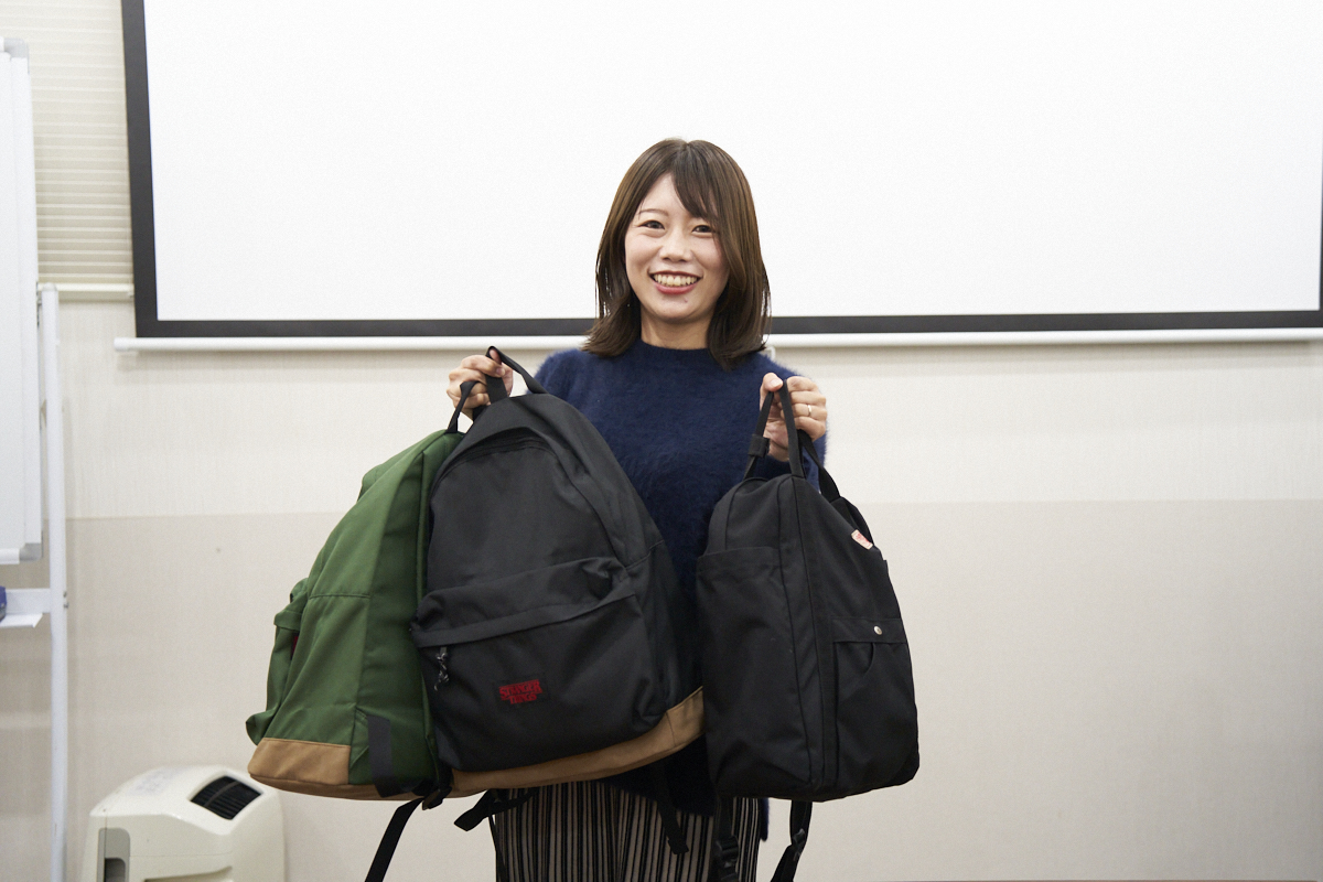 菱山さんが3つの防災バッグを持ちながら笑顔で立っています。バッグは異なるデザインで、それぞれの用途に合わせて選ばれています。