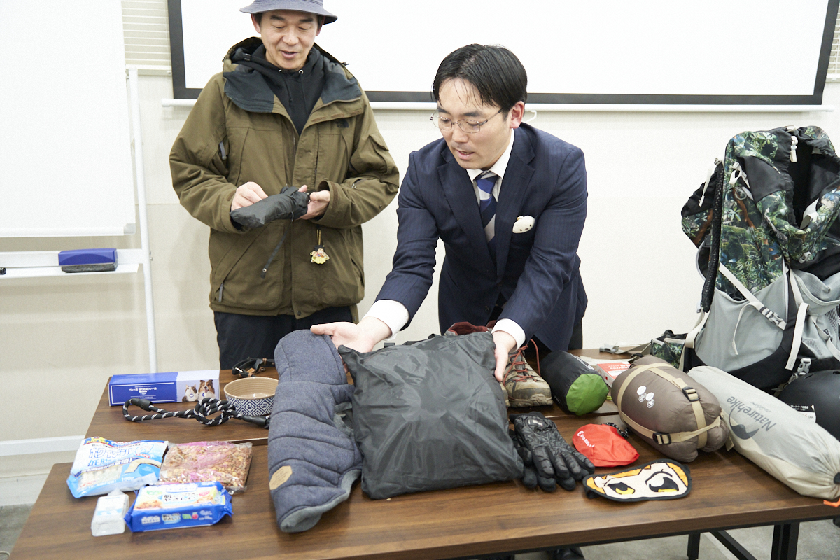 テーブルに並べられたアウトドア用品を、スーツ姿の男性が確認しています。隣には帽子をかぶった男性が立っています。