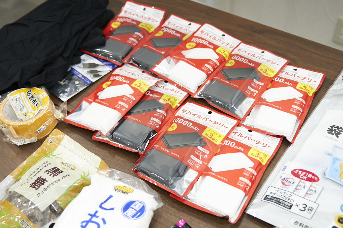 テーブルの上に、複数のモバイルバッテリー、粘着テープ、黒糖、塩などの防災用品が整然と並べられています。