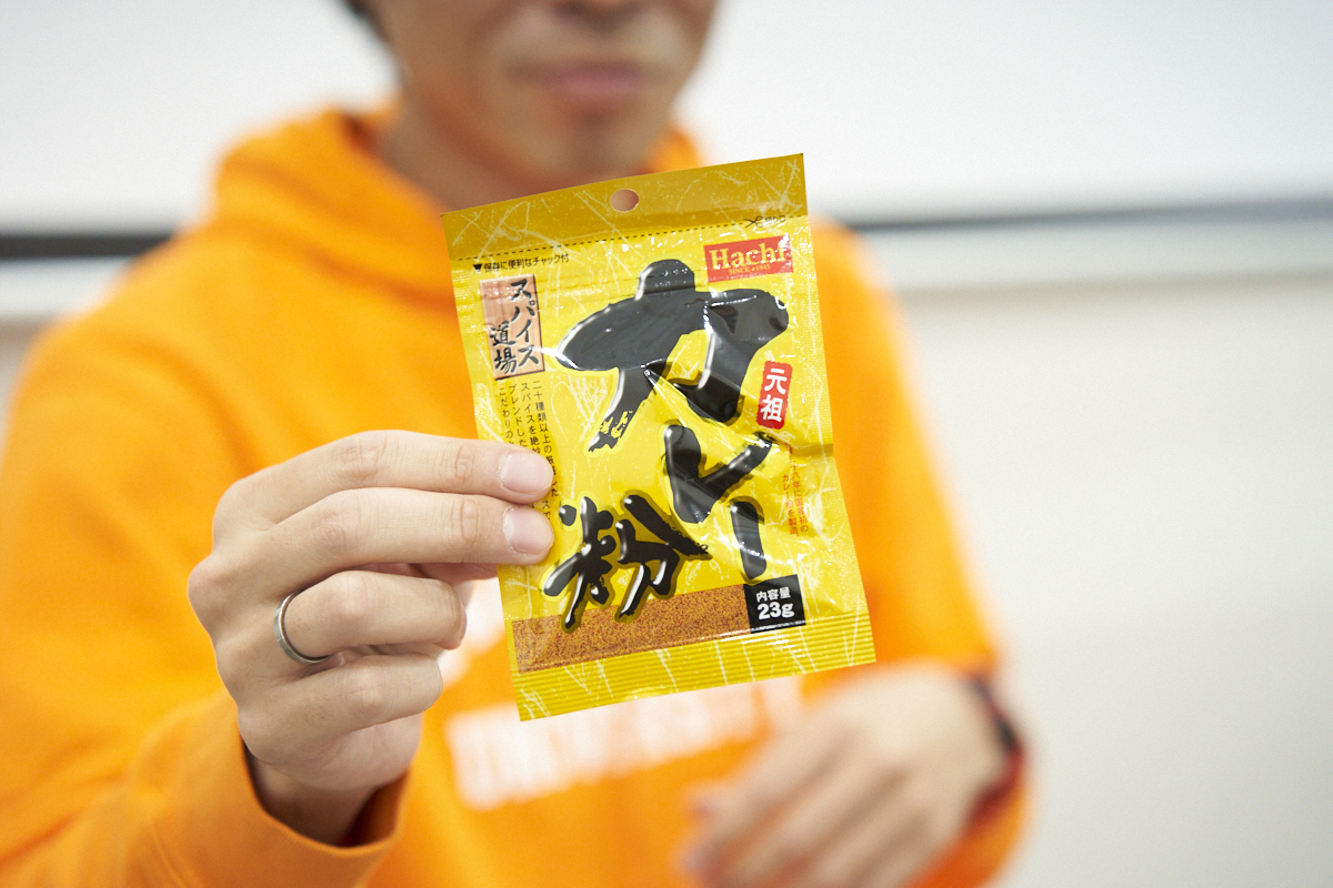 オレンジ色の服を着た地主さんが、黄色いパッケージの「カレー粉」を手に持っている様子です。